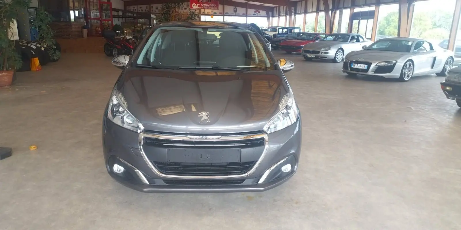 Vue avant d'une Peugeot 208 gris métal 2023 exposée en intérieur avec calandre chromée et phares allumés.