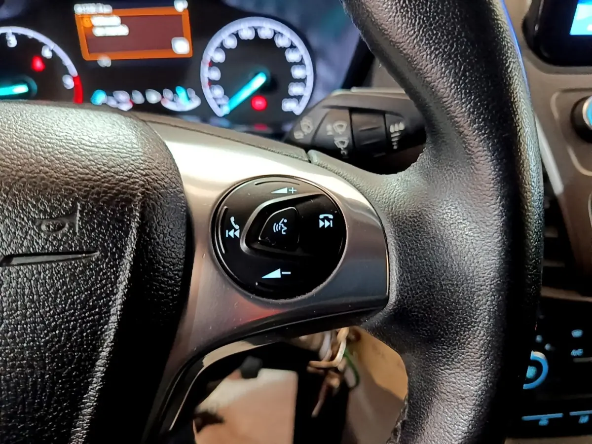 Gros plan sur le volant noir du Ford Transit Fourgon 2022 avec commandes vocales et tableau de bord éclairé en arrière-plan.