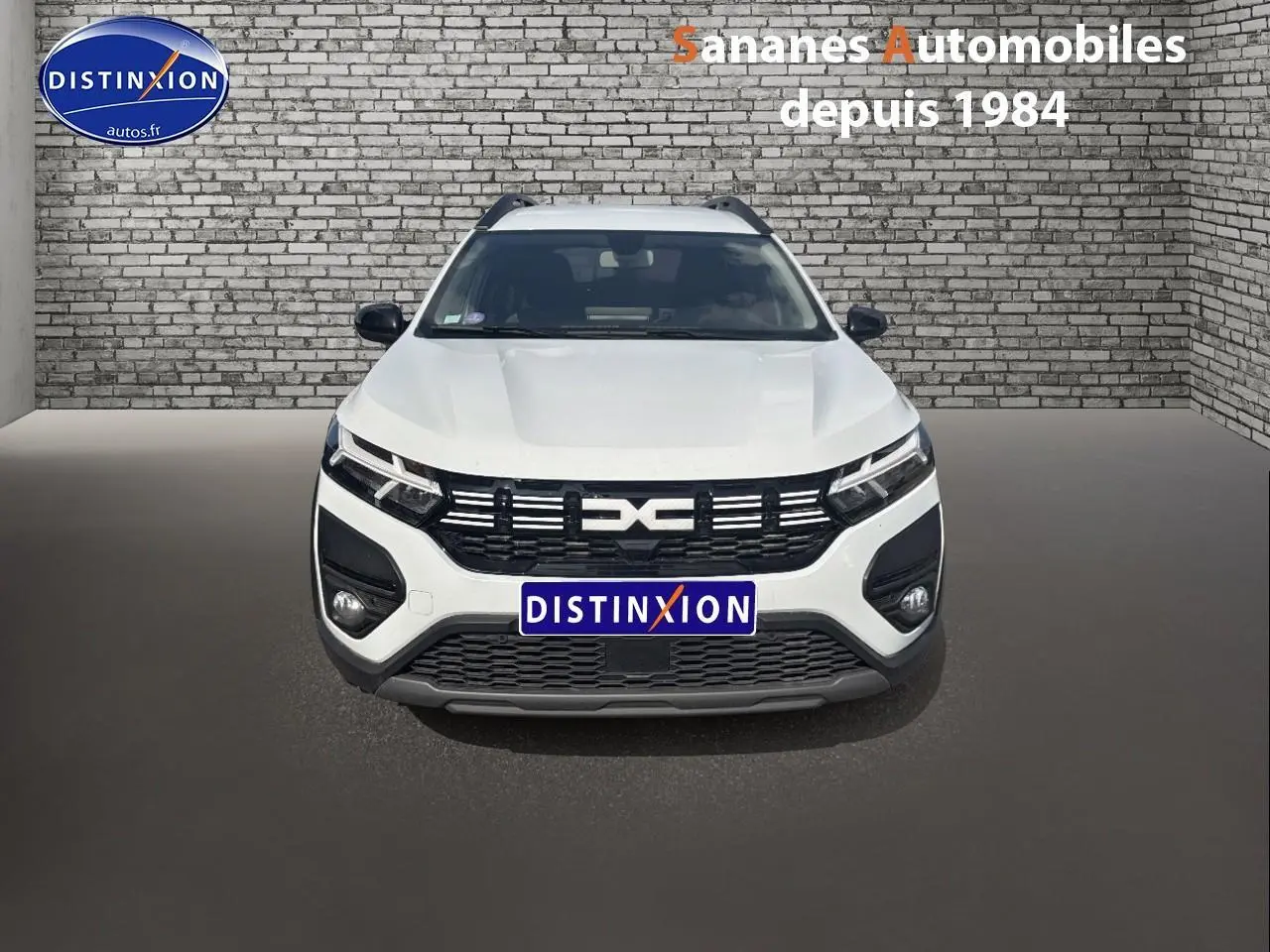 Vue frontale d'un Dacia Jogger blanc avec calandre noire et logo distinctif, stationné devant un mur en briques grises.