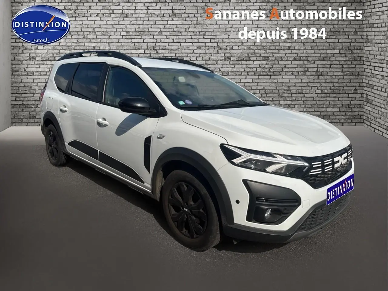 Dacia Jogger blanc vue 3/4 avant gauche avec jantes noires et barres de toit sur fond de mur en briques.