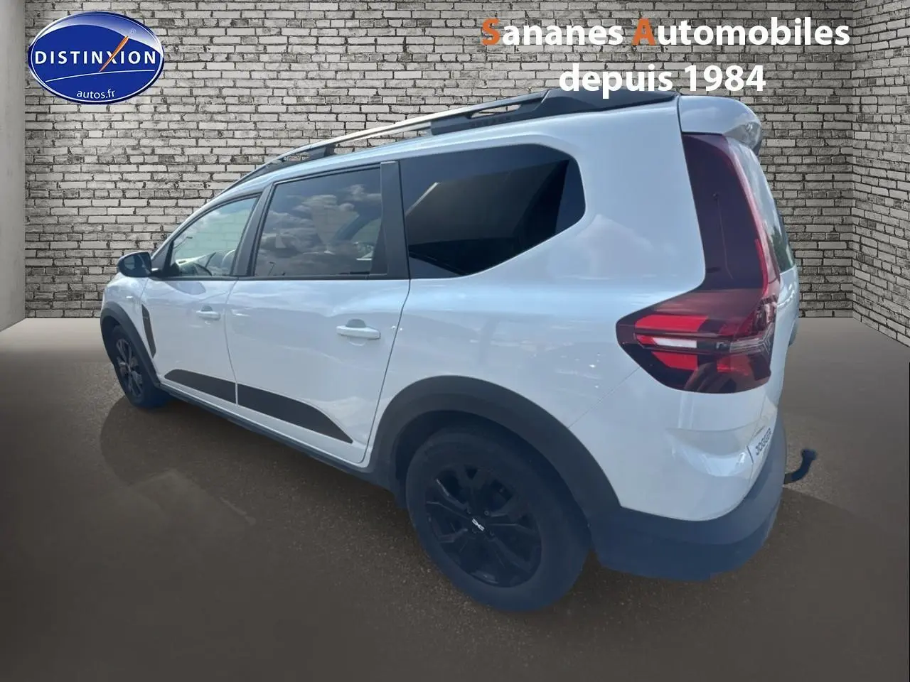 Vue 3/4 arrière droite d'un Dacia Jogger blanc avec jantes noires et attelage fixe sur fond brique.