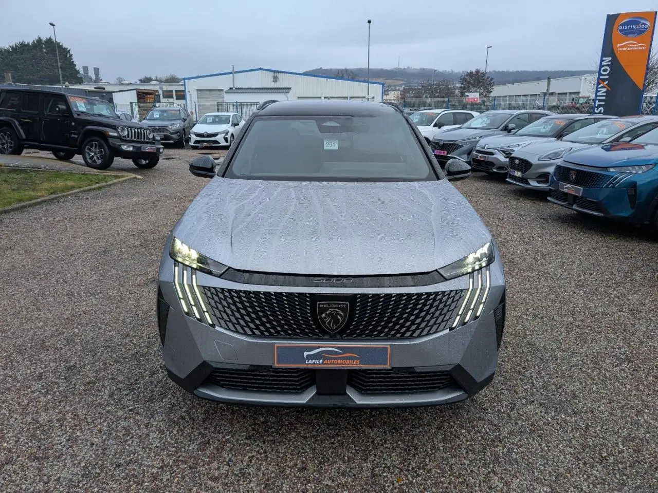 Vue avant d'un Peugeot 5008 gris métallisé 2025 avec calandre noire et feux LED en forme de griffes allumés.