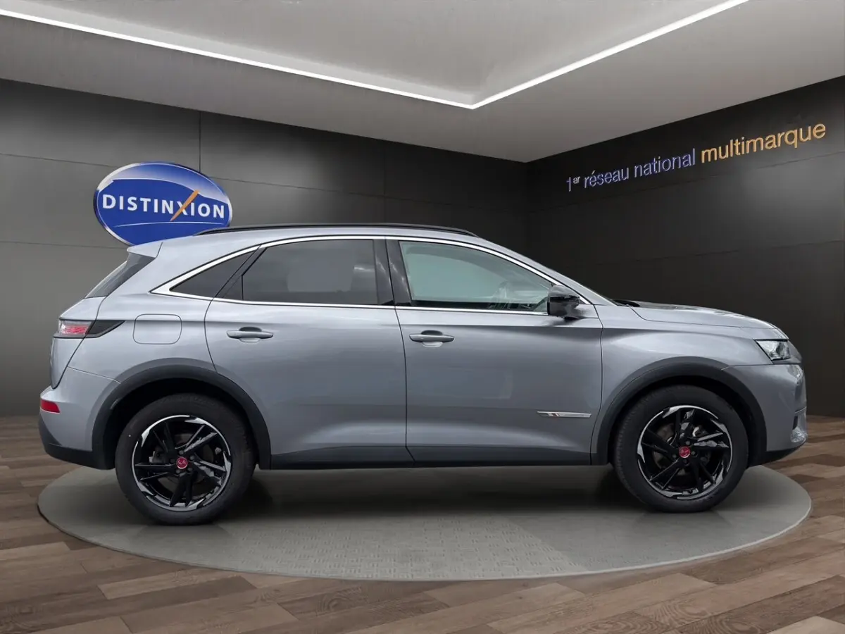 Vue de profil côté droit d'un DS7 Crossback gris clair avec jantes noires et détails chromés sur fond intérieur showroom.