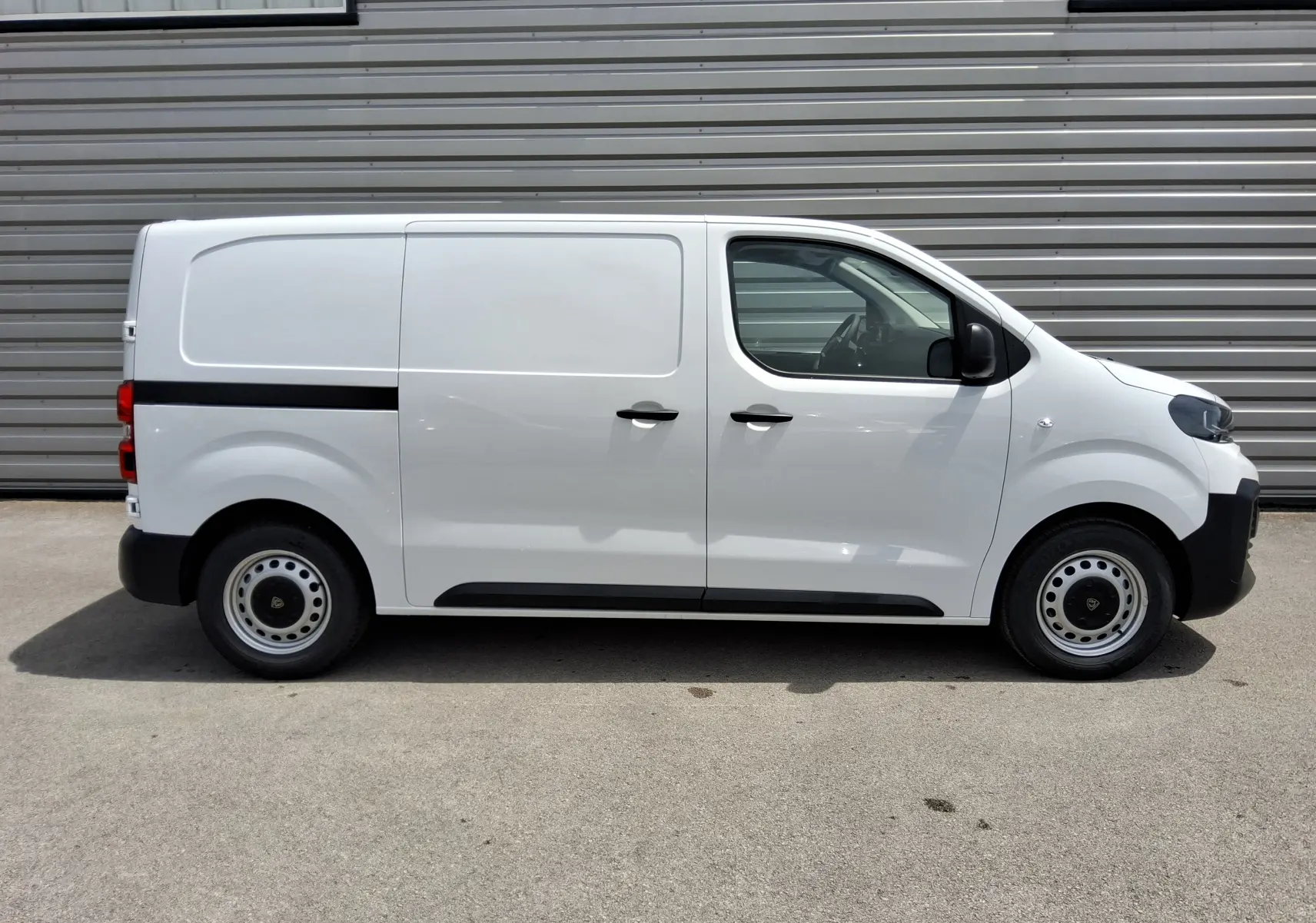 Vue latérale droite d’un Peugeot Expert Fourgon blanc 2025 avec bande noire et roues acier, stationné devant un mur métallique.