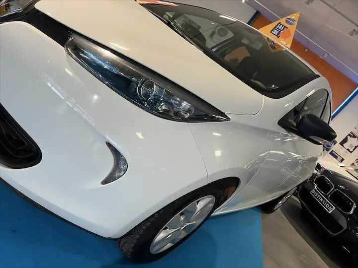 Vue 3/4 avant gauche d'une Renault ZOE blanche glacier électrique en showroom avec détails de phares et jantes visibles