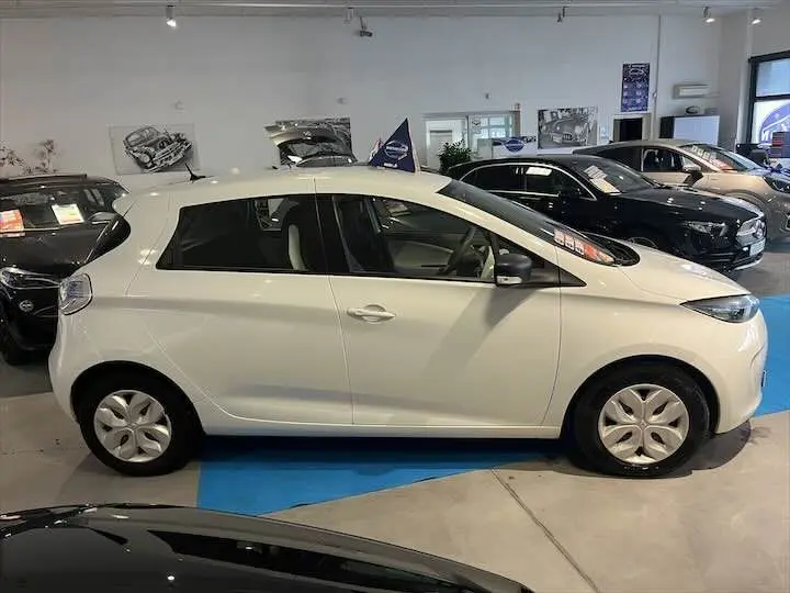 Profil latéral droit d'une Renault ZOE R90 LIFE blanc glacier exposée en showroom avec jantes acier et vitres teintées.