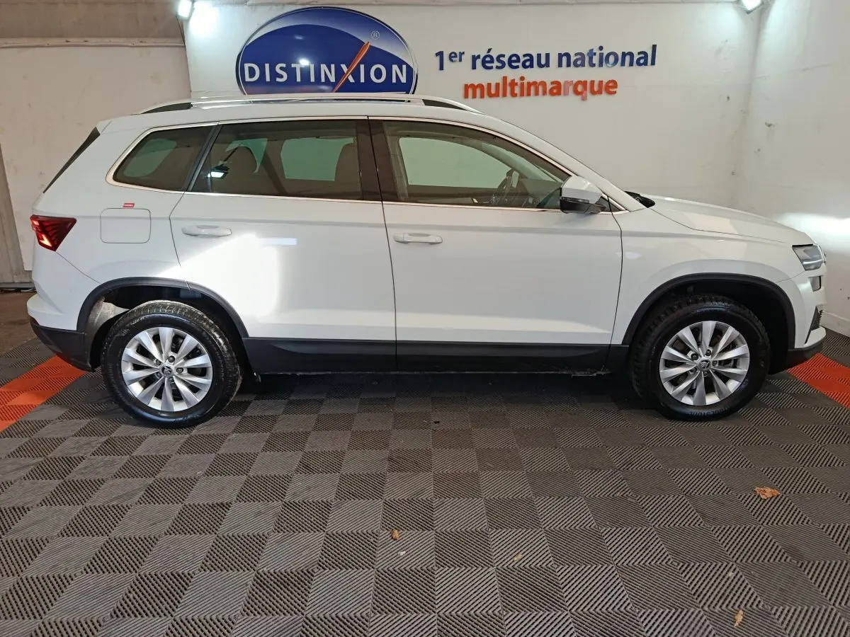 Vue profil droit d'un Skoda Karoq blanc 2024 avec jantes alliage et rails de toit noirs en intérieur showroom.