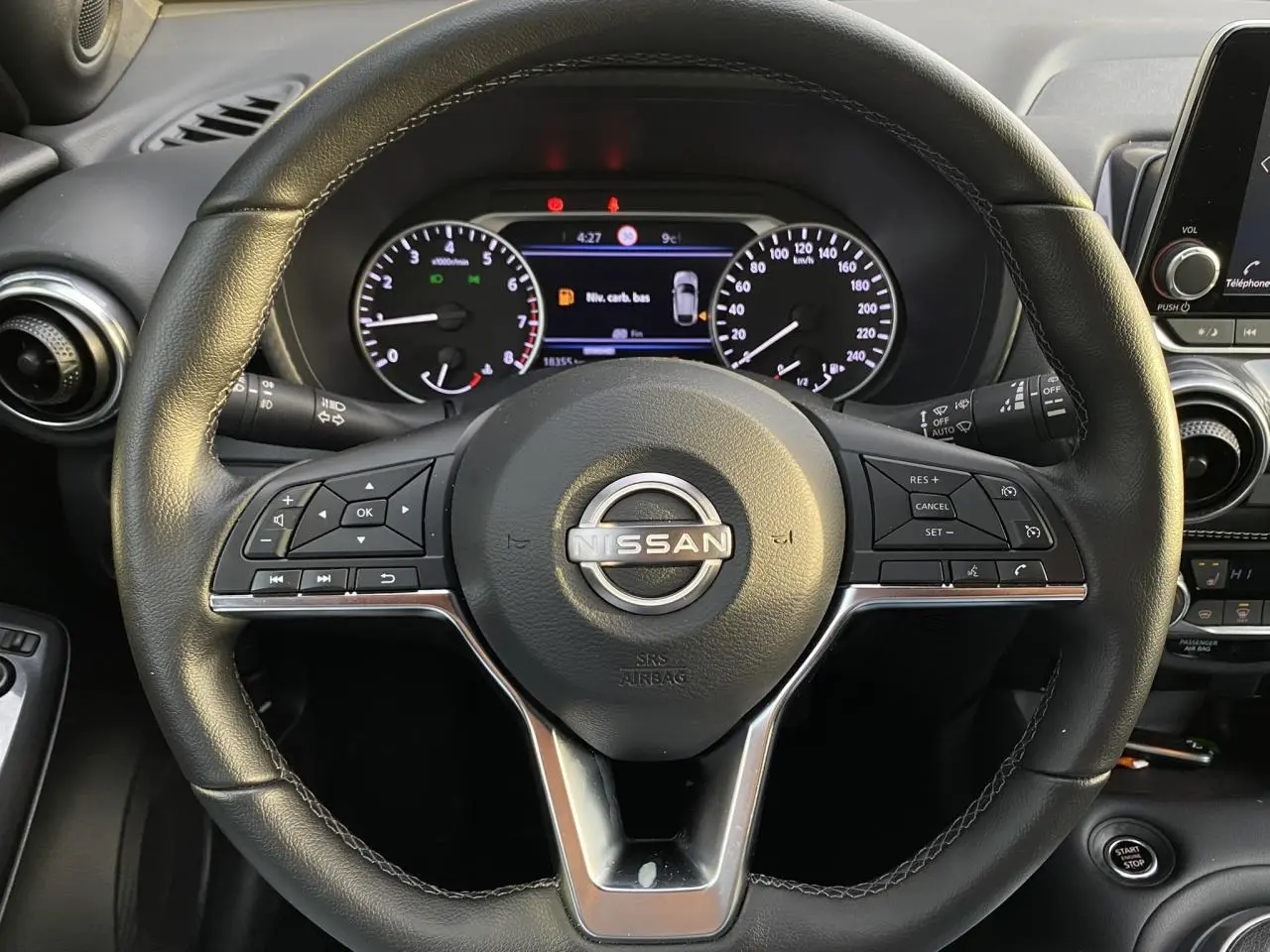 Vue rapprochée du volant noir du Nissan Juke 2024 avec commandes intégrées et tableau de bord numérique en arrière-plan.
