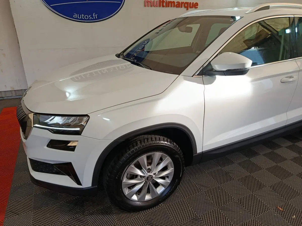 Vue 3/4 avant droit d’un Skoda Karoq blanc 2024 avec jantes alliage et rétroviseurs blancs sur fond intérieur.