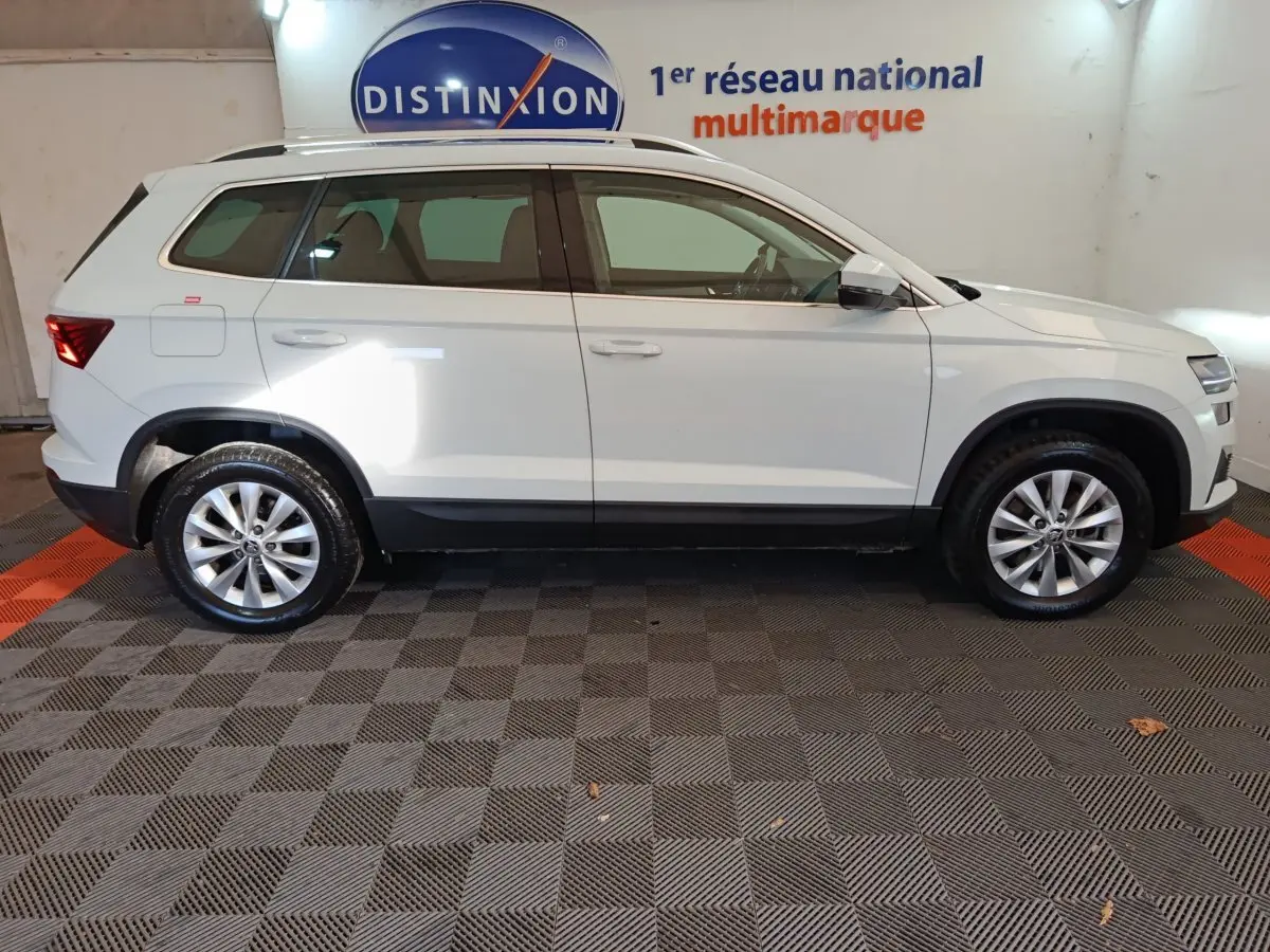 Vue profil côté gauche d'un Skoda Karoq blanc 2024 sur sol damier, avec jantes alliage et rétroviseurs électriques.