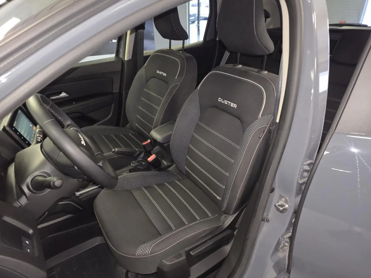 Vue intérieure côté conducteur du Dacia Duster gris 2024, sièges noirs avec surpiqûres blanches et logo Duster brodé.