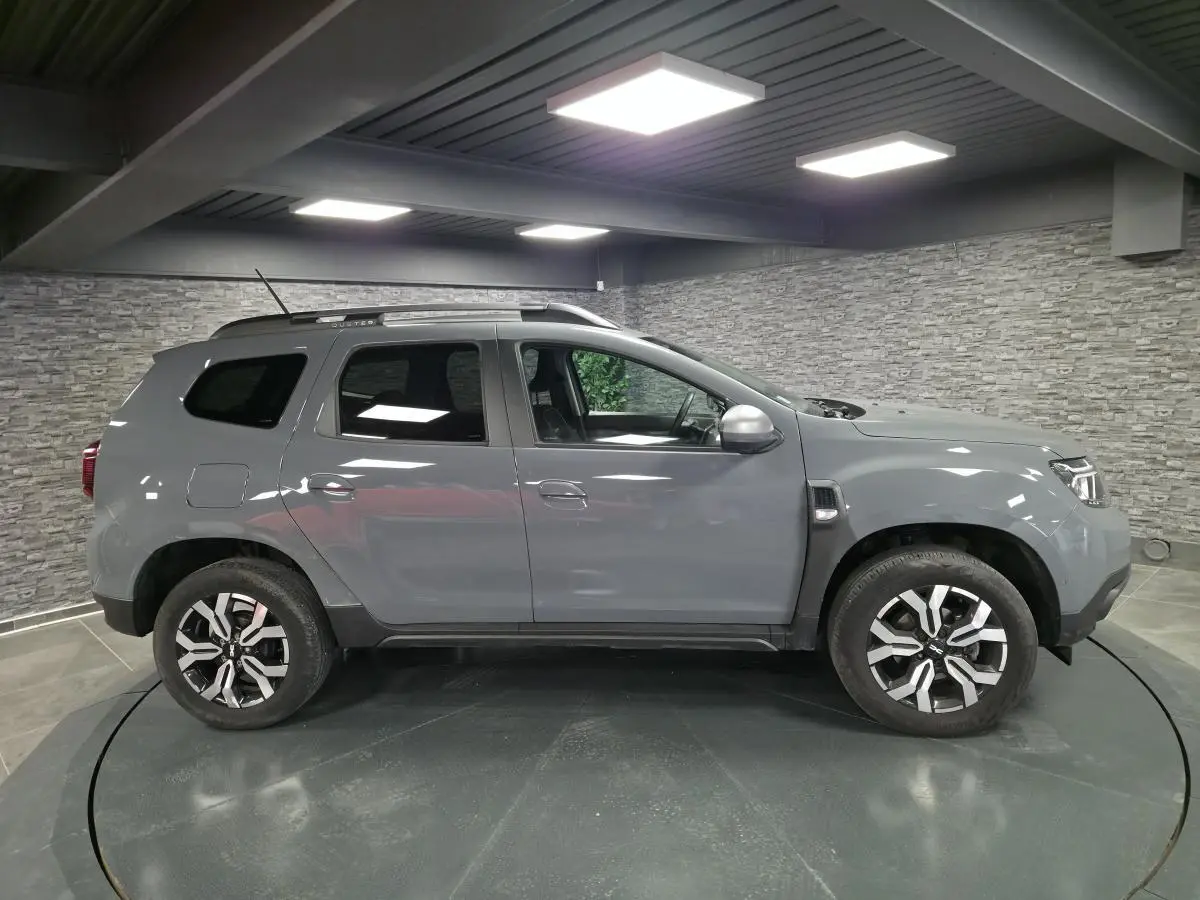 Vue de profil droit du Dacia Duster 2024 gris urbain avec jantes alliage 17 pouces et barres de toit gris.