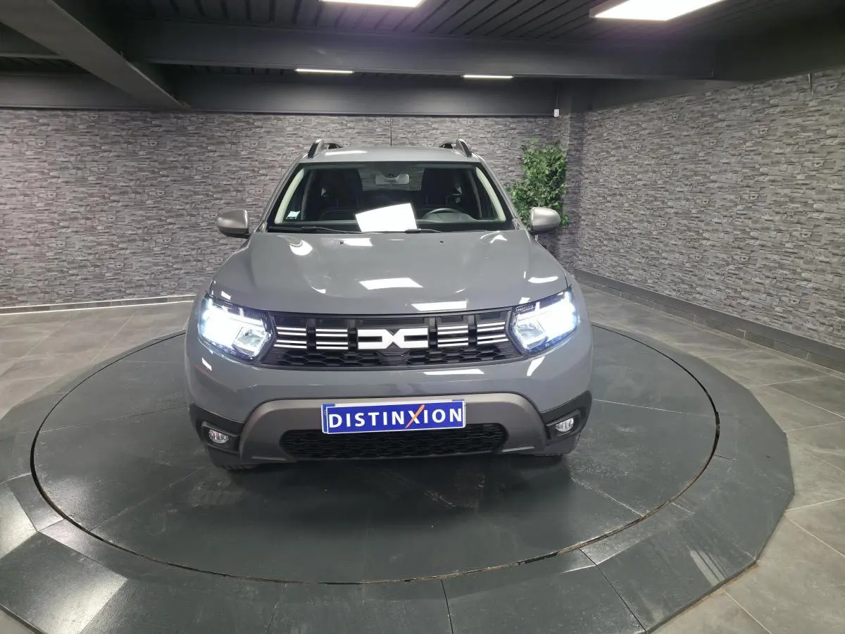 Vue avant d'un Dacia Duster 2024 gris urbain avec calandre noire et phares allumés en intérieur.
