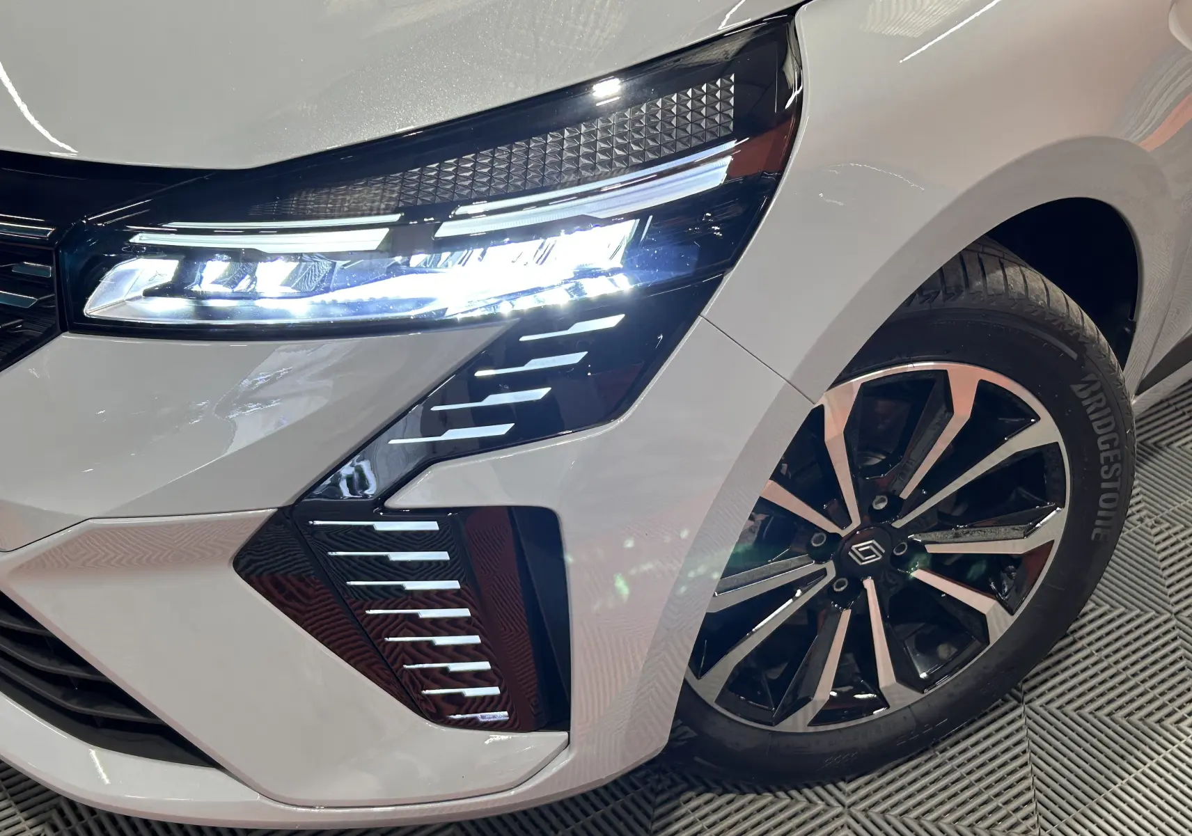 Gros plan sur l'avant droit gris Rafale de la Renault Clio V E-Tech avec phares LED allumés et jante noire brillante.