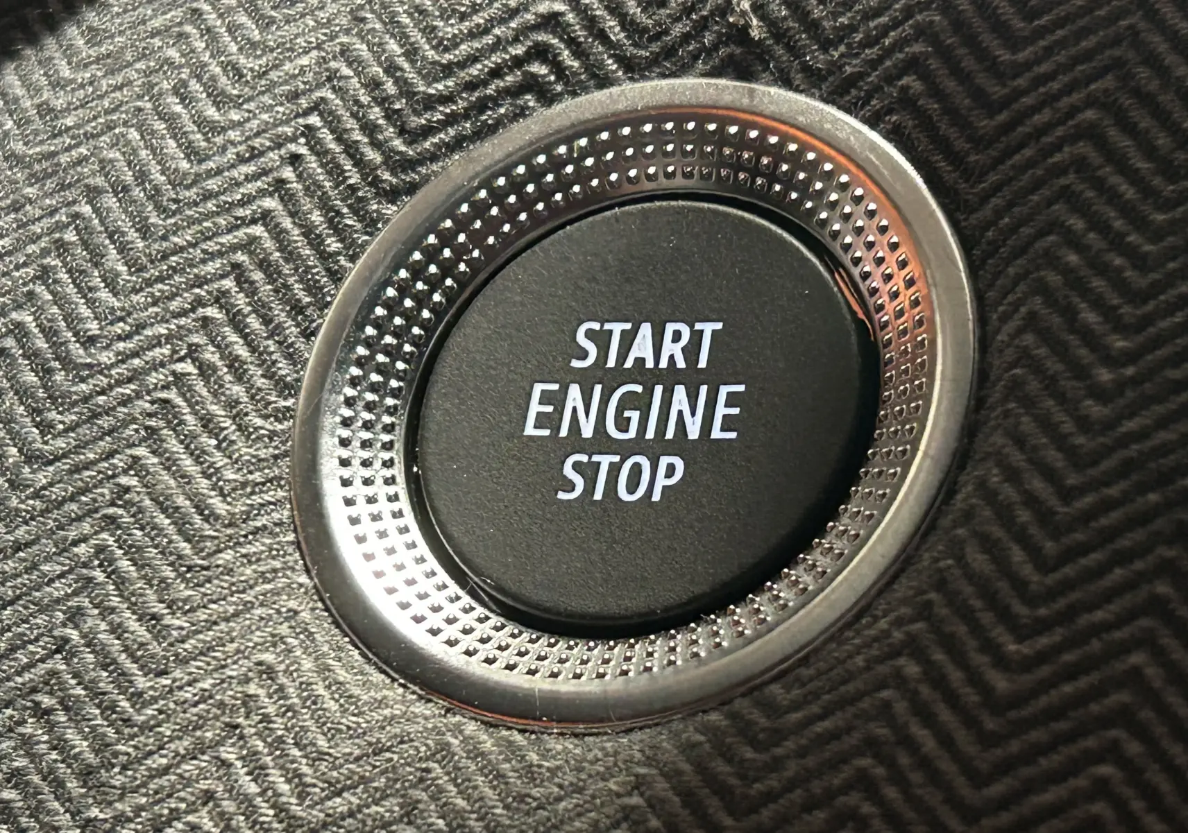 Gros plan sur le bouton Start Engine Stop entouré d’un cerclage métallique, sur un revêtement tissu gris foncé à motif chevrons.