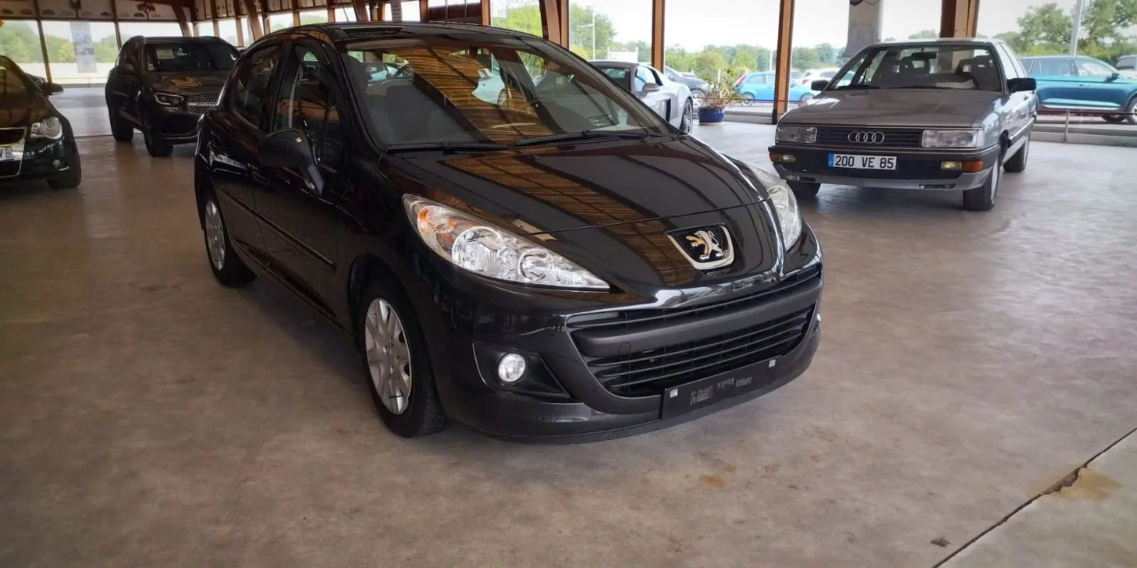 Peugeot 207+ noire vue 3/4 avant droit dans un garage, mettant en valeur ses phares et calandre distinctifs.