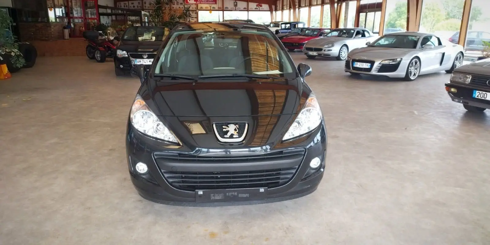 Vue avant d'une Peugeot 207+ noire de 2013, avec phares allumés, stationnée dans un showroom lumineux.