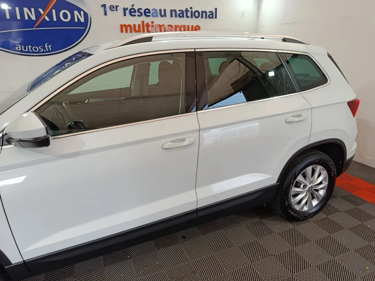Vue latérale droite d'un Skoda Karoq blanc 2024 avec jantes alliage et barres de toit noires visibles en intérieur showroom.
