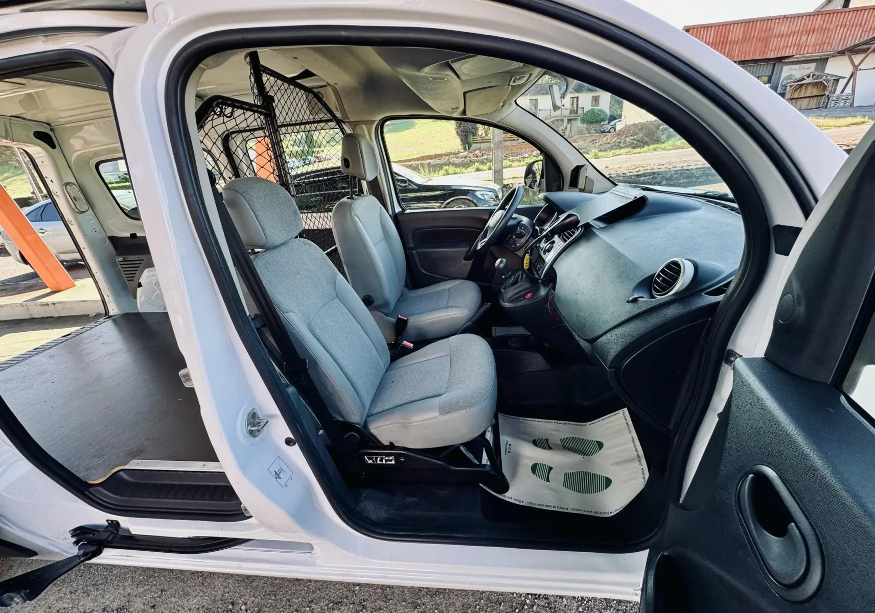 Vue intérieure côté conducteur du Renault Kangoo Maxi blanc, montrant les sièges gris et la cabine utilitaire.