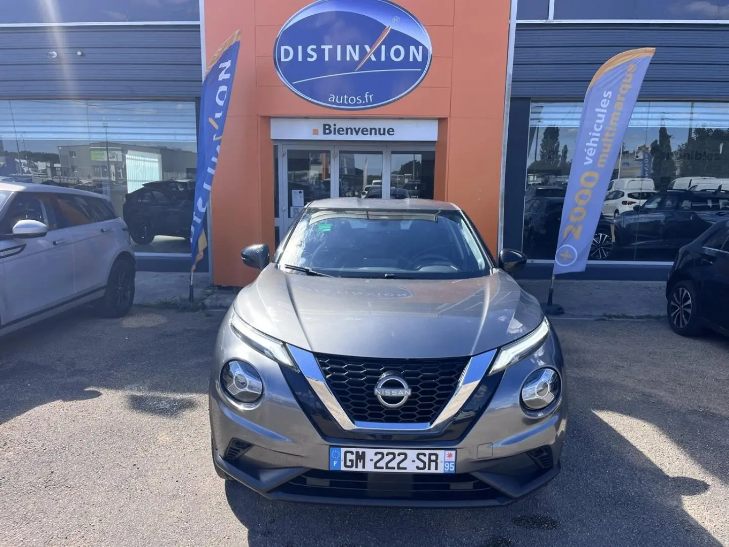 Nissan Juke 2023 gris argile vue de face devant une concession avec drapeaux publicitaires.