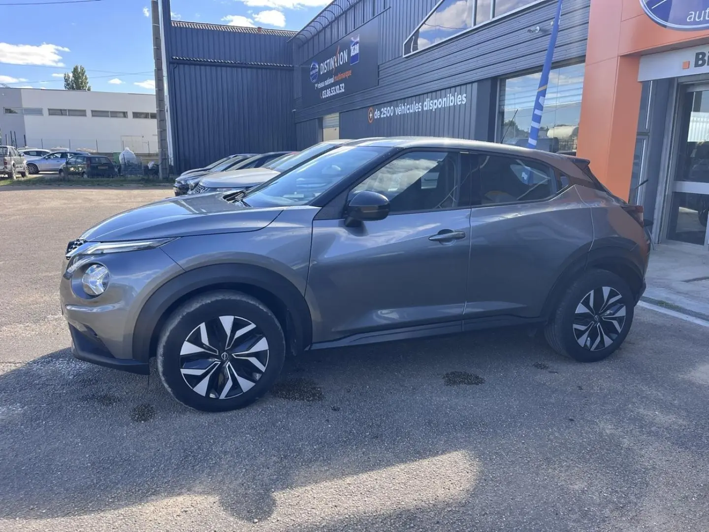 Profil droit d'un Nissan Juke 2023 gris argile avec jantes bi-ton et poignées ton carrosserie, stationné devant un bâtiment.