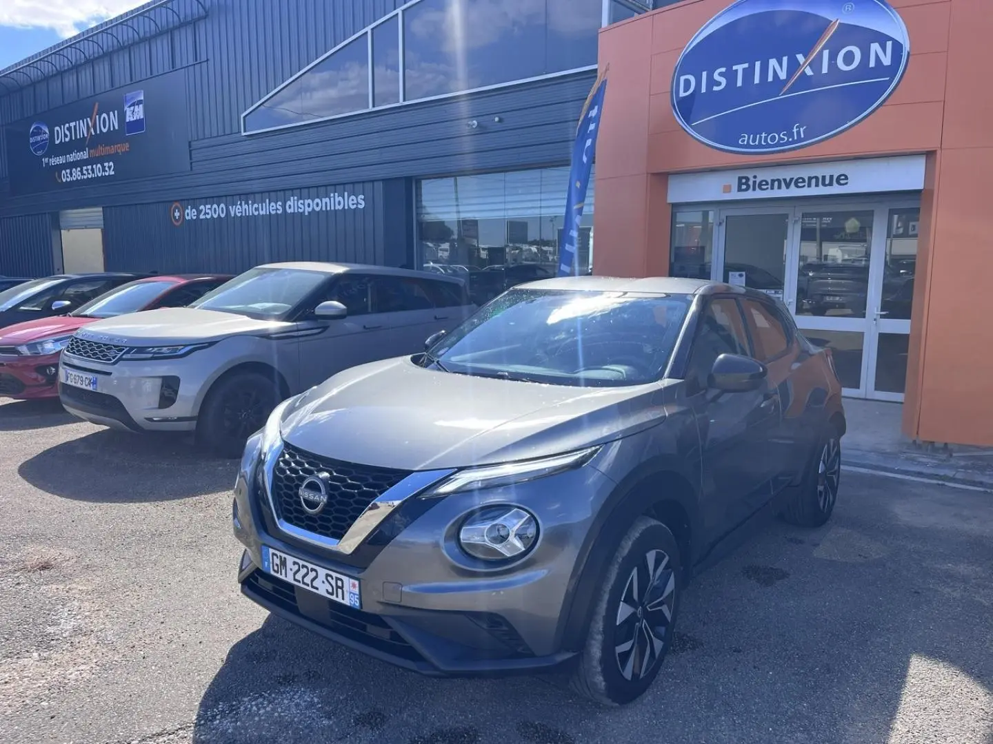 Nissan Juke gris argile vue 3/4 avant droit, avec jantes bi-ton et calandre chromée, stationné devant un concessionnaire.
