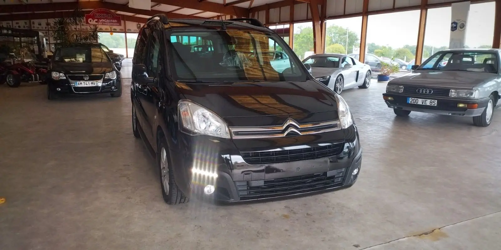 Vue 3/4 avant d'un Citroën Berlingo noir 2016, avec phares allumés, dans un showroom lumineux.