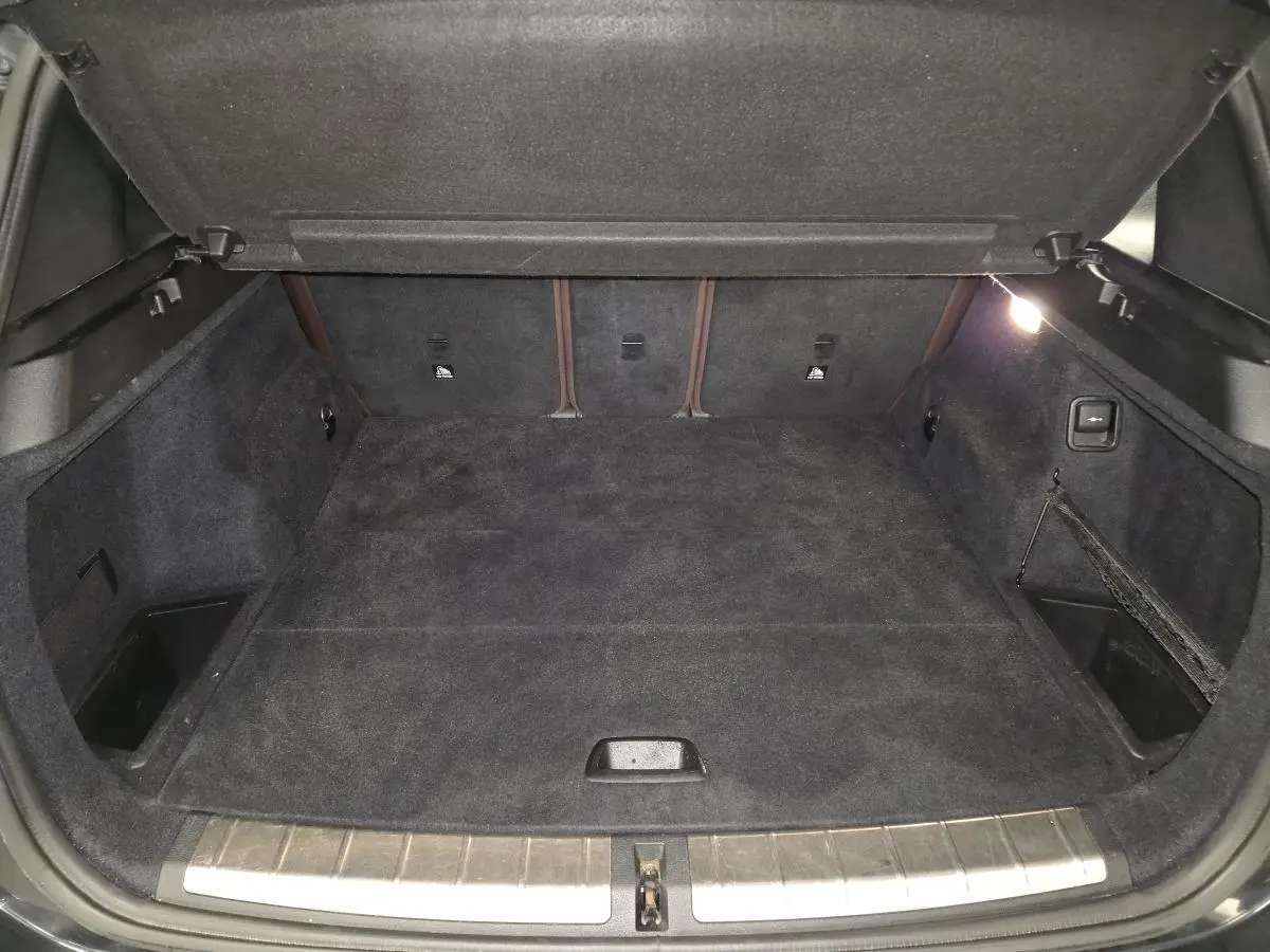 Coffre ouvert vu de face, intérieur noir avec tapis et rangements latéraux, hayon électrique visible sur BMW X1 noir.
