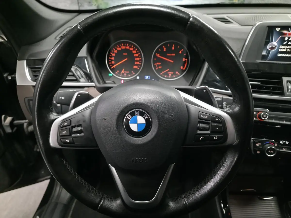 Vue rapprochée du volant cuir noir multifonctions d'une BMW X1 xDrive 25d 2016 avec tableau de bord et écran central visibles.