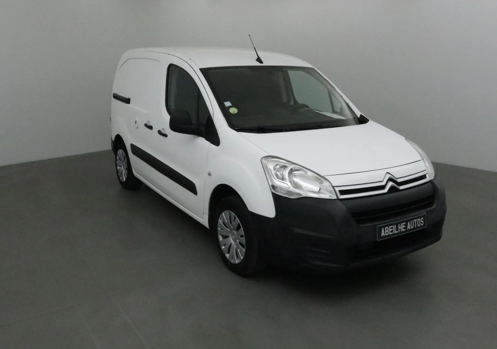 Vue 3/4 avant gauche d'un utilitaire Citroën Berlingo blanc avec pare-chocs noirs dans un studio gris.