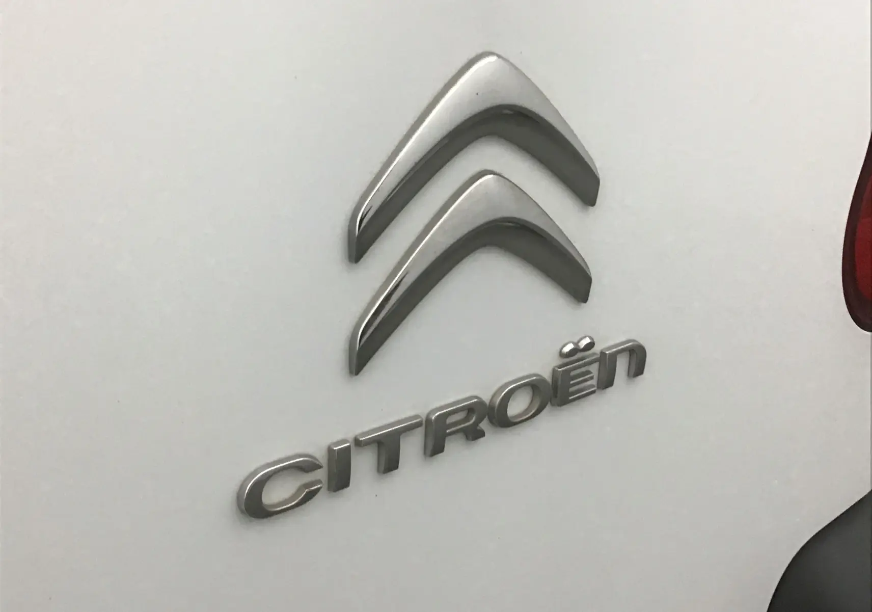 Gros plan sur le logo argenté Citroën sur la porte arrière blanche d'un Berlingo 2018, avec un feu arrière partiellement visible.
