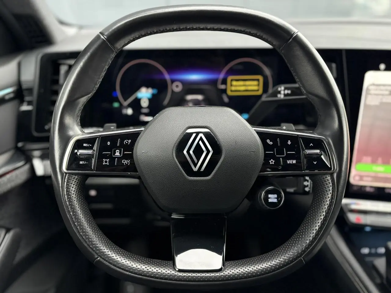 Vue rapprochée du volant noir perforé avec logo Renault au centre, tableau de bord numérique en arrière-plan flou.