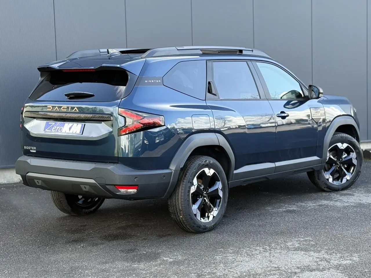 Vue 3/4 arrière droite du Dacia Bigster bleu indigo avec jantes alliage noires et hayon électrique visible.