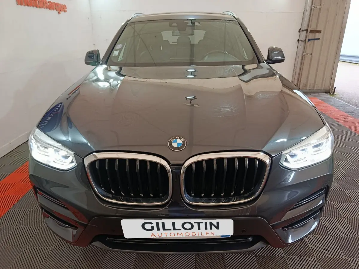 Vue avant d'une BMW X3 xDrive30e gris foncé avec calandre double haricot et phares LED allumés dans un garage.