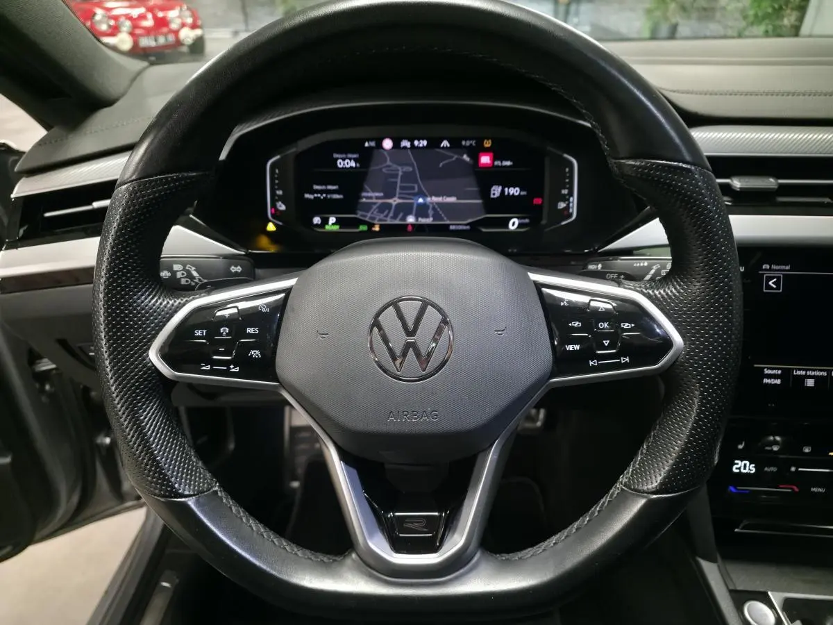 Vue frontale du volant noir perforé avec logo Volkswagen et tableau de bord digital affichant la navigation du Volkswagen Arteon Shooting Brake gris.