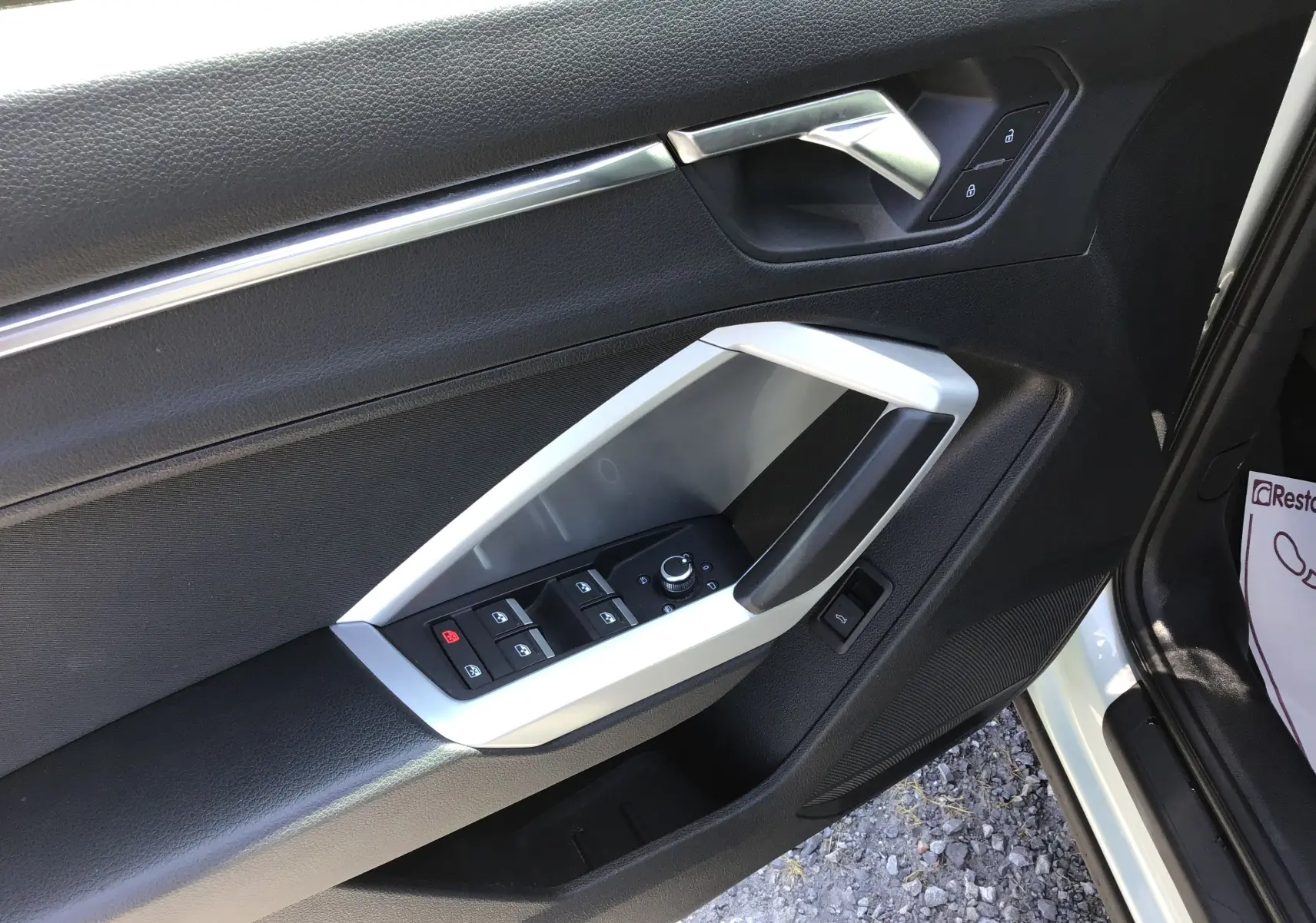 Vue rapprochée de la porte avant gauche de l'Audi Q3 Sportback gris clair, avec commandes électriques et poignée argentée.