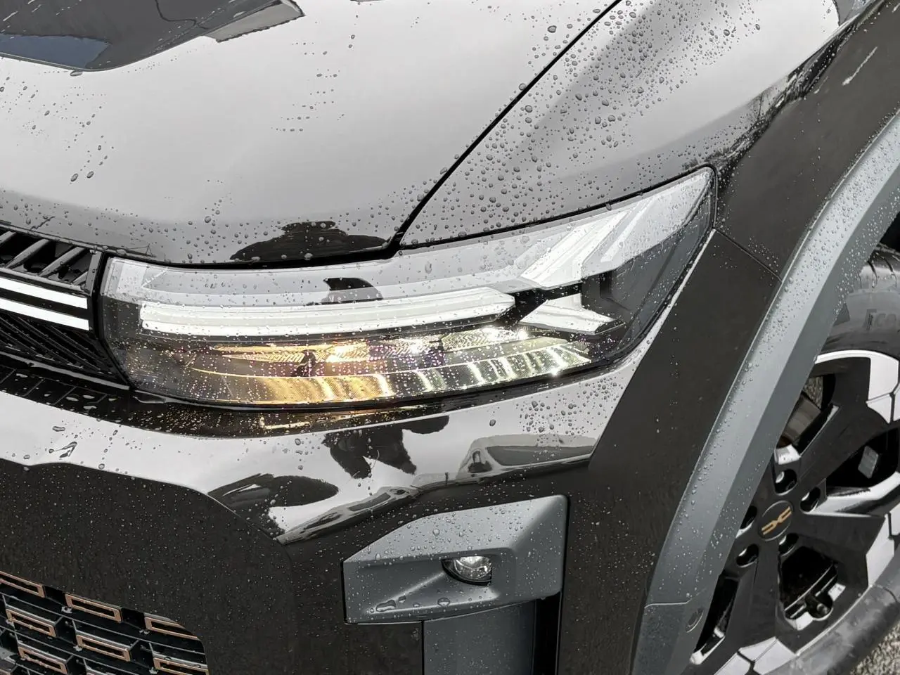 Gros plan sur le phare avant droit et la jante noire diamantée du Dacia Bigster noir nacré sous la pluie.