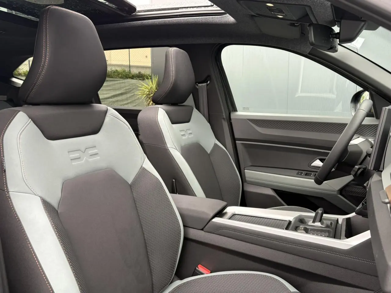 Vue intérieure côté droit du Dacia Bigster 2026, sièges bicolores gris clair et foncé avec logo embossé, toit panoramique visible.
