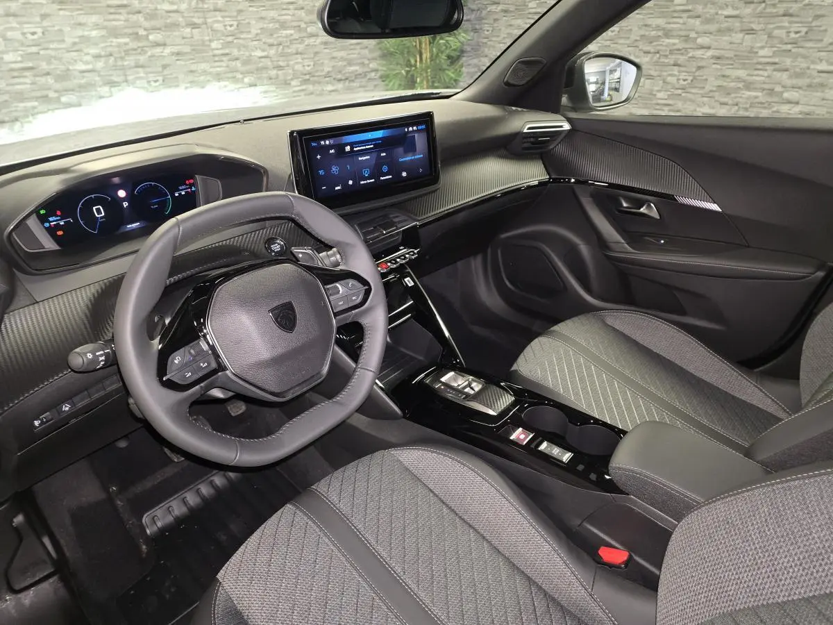 Vue intérieure avant droite du Peugeot 2008 1.2i Hybrid 2025, avec volant compact, tableau de bord numérique et sellerie tissu gris.