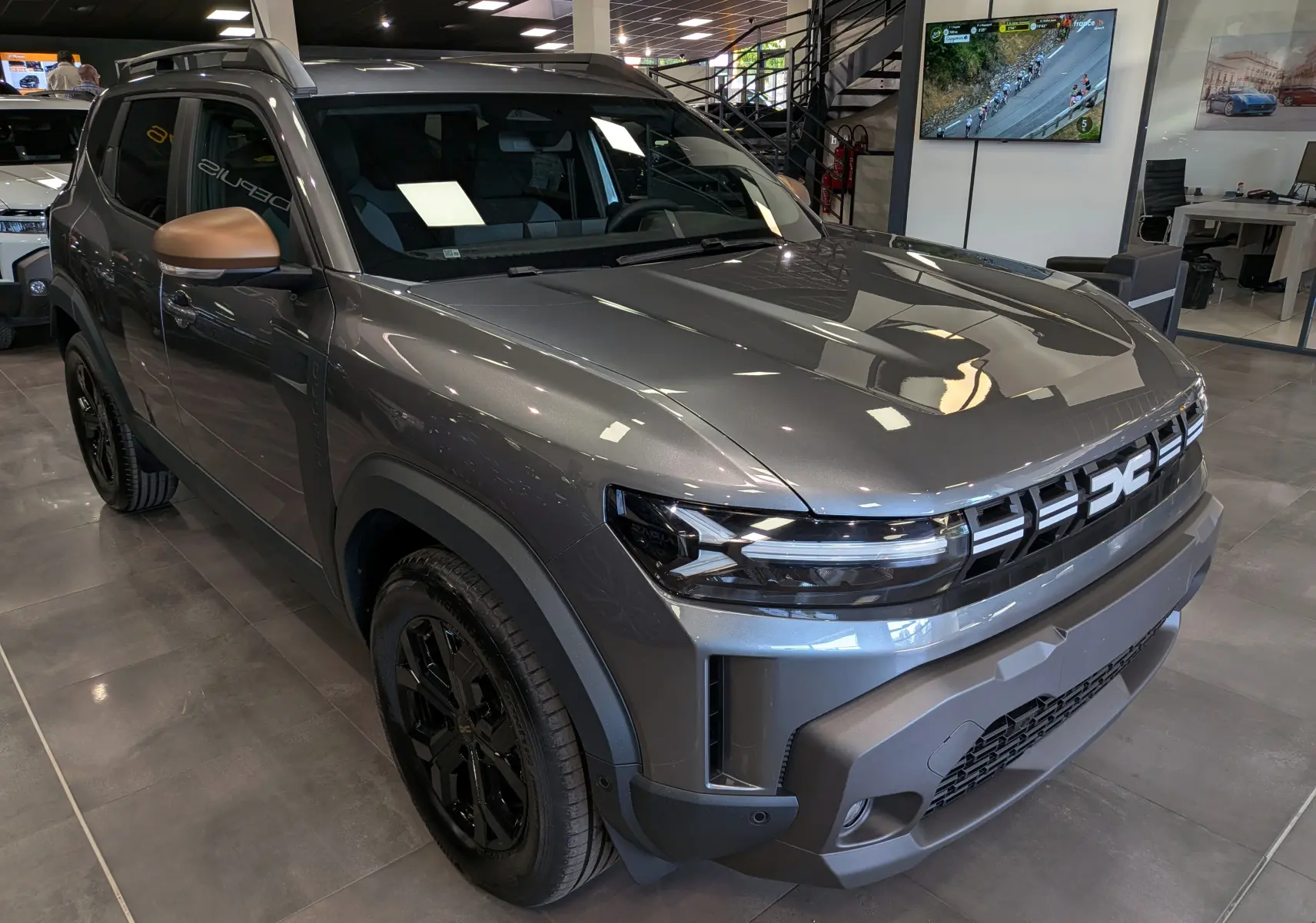 Dacia Duster 2026 gris schiste en vue 3/4 avant droit, avec rétroviseurs bronze et jantes noires brillantes.