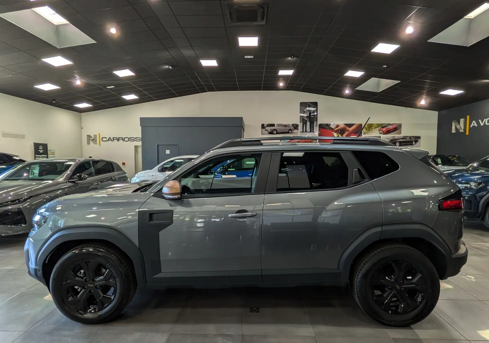 Profil droit du Dacia Duster 2026 gris schiste avec jantes noires et rétroviseurs couleur bronze en showroom.