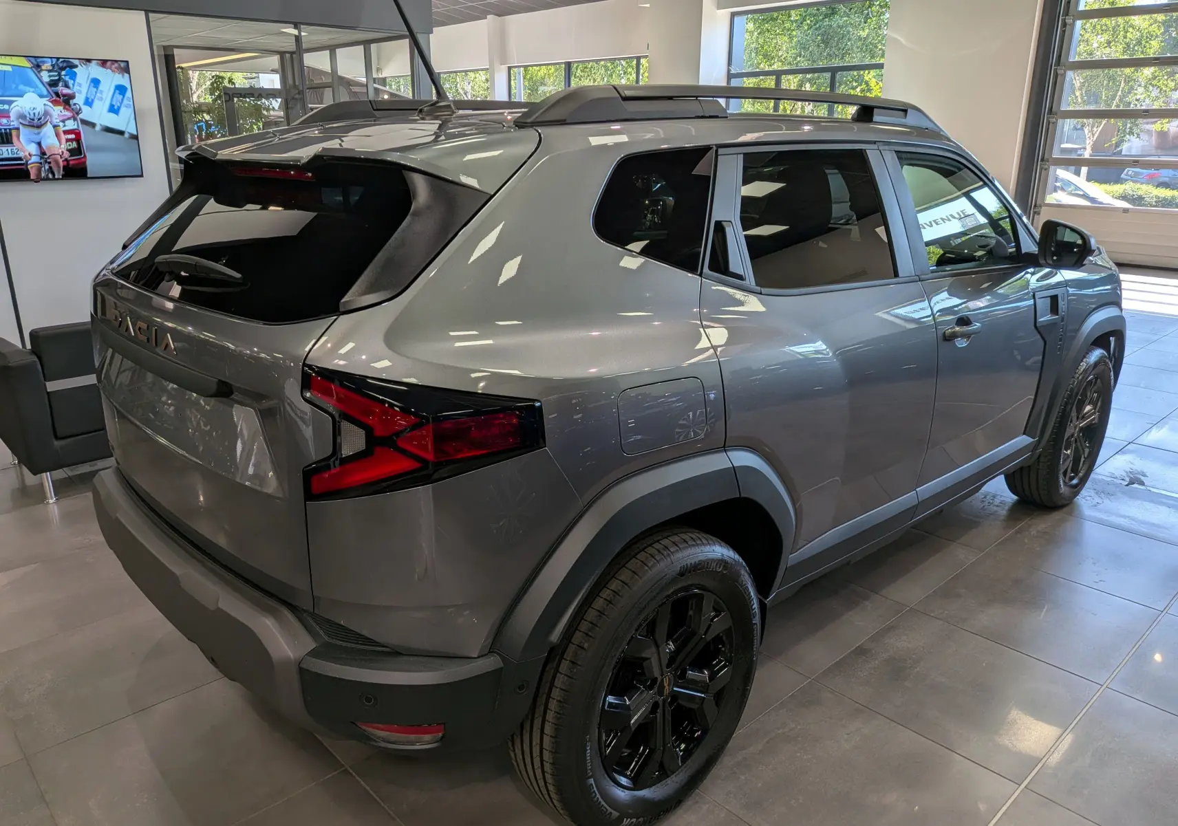 Vue 3/4 arrière droit du Dacia Duster 2026 gris schiste avec jantes noires et barres de toit en showroom.