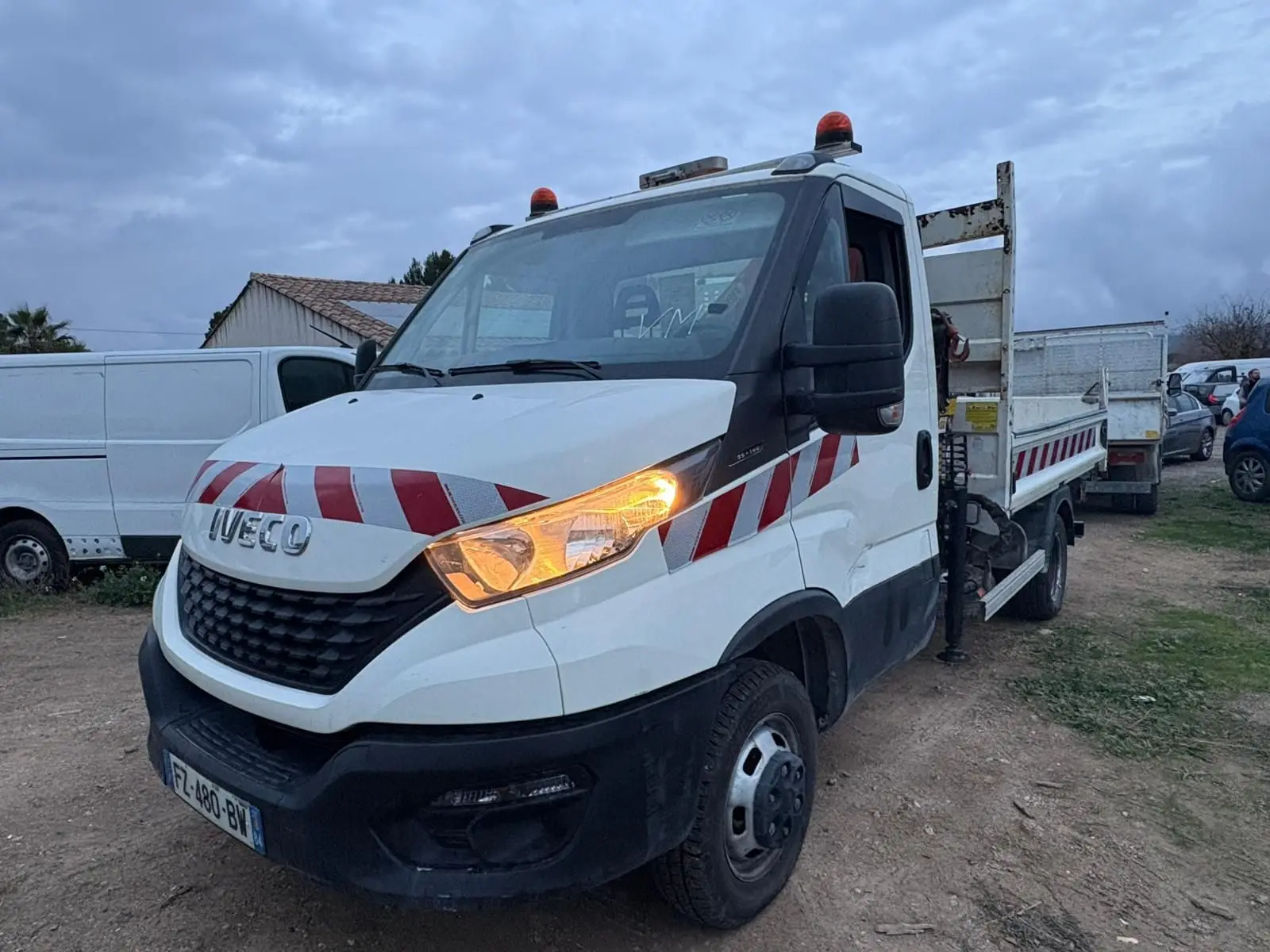 IVECO Daily 35C14 blanc en 3/4 avant droit, avec benne, grue Palfinger et feux allumés.