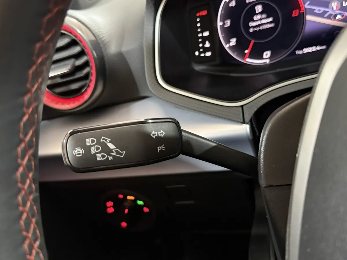 Gros plan sur la manette de clignotants noire avec marquages blancs, volant cuir rouge et tableau de bord digital d'une SEAT Ibiza blanche.