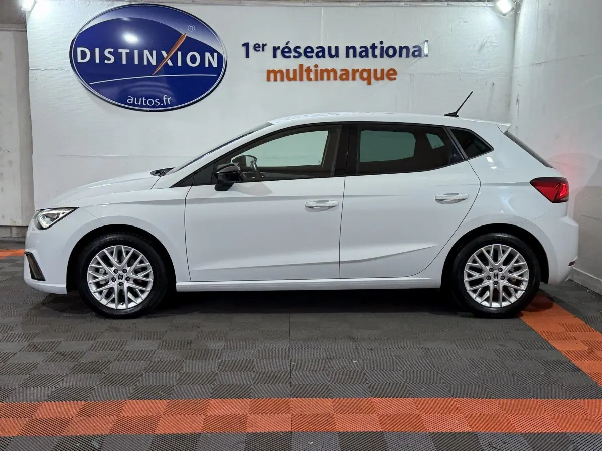 Profil droit d'une SEAT Ibiza blanche 2024, compacte, avec jantes alliage et feux arrière allumés en intérieur showroom.
