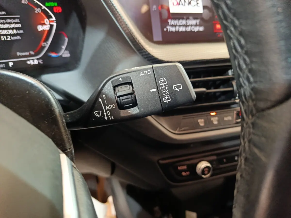 Gros plan sur la commande d’essuie-glace à gauche du volant d’une BMW Série 1 noire, avec tableau de bord numérique en arrière-plan.