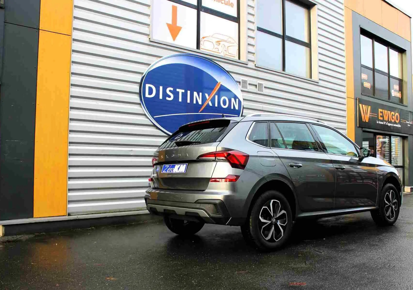 Vue 3/4 arrière droite d'un Skoda Kamiq gris graphite 2025 garé devant un bâtiment commercial.