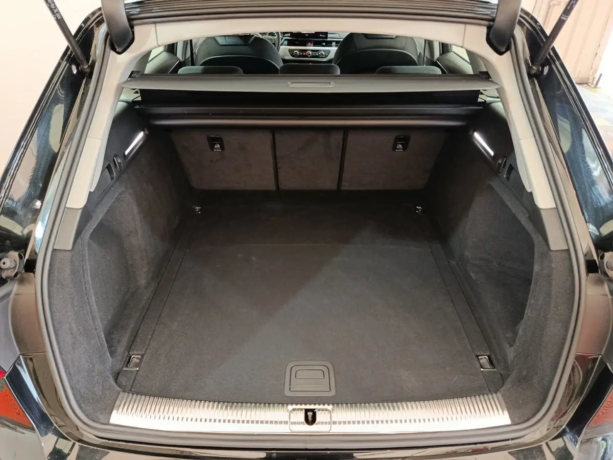 Coffre ouvert vu de l'arrière d'une Audi A4 noire 2024, intérieur spacieux avec tapis noir et éclairage latéral.