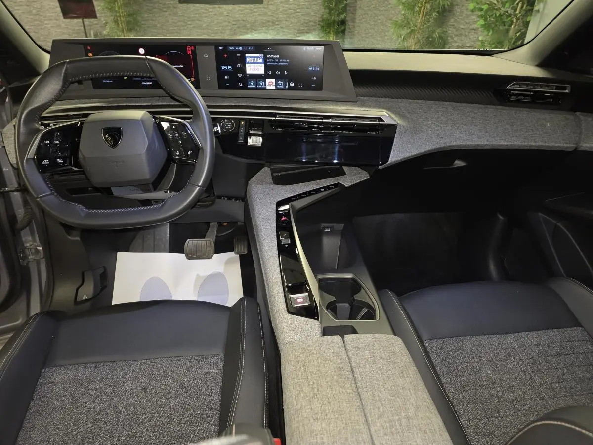 Vue intérieure avant du Peugeot 3008 Hybrid 2025 avec tableau de bord tissu gris, volant compact cuir noir et double écran HD flottant.