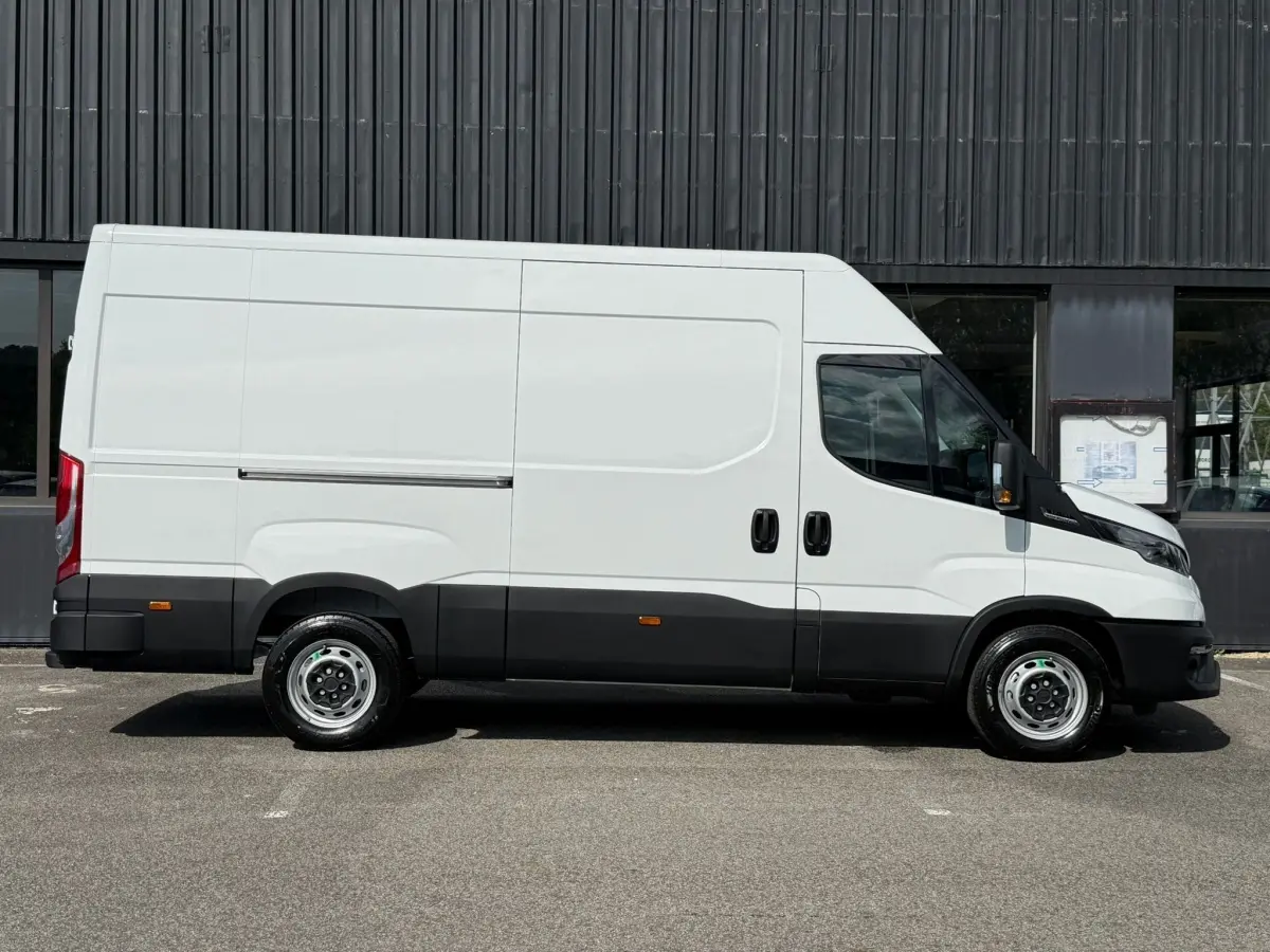 Vue de profil côté droit d’un utilitaire Iveco Daily blanc avec bas de caisse noir, stationné devant un mur sombre.